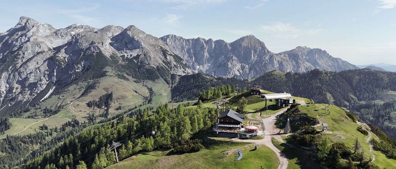 Luftaufnahme einer Berglandschaft mit einem Skigebiet. Die Berge sind von grünen Bäumen umgeben und haben einen klaren blauen Himmel. Ein Skilift ist in der Ferne sichtbar.