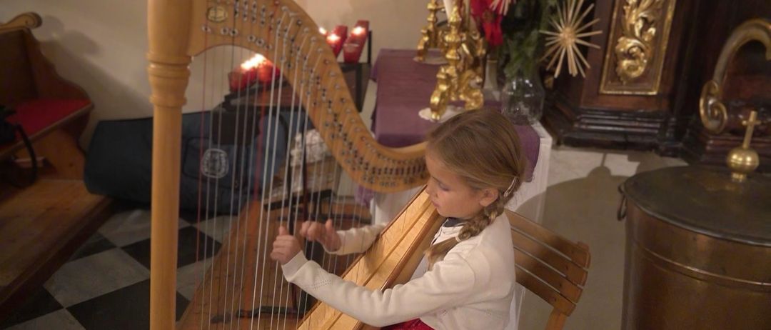 Ein kleines Mädchen spielt eine Harfe in einem Raum mit Kerzen und einer dekorativen Pflanze. Sie trägt ein weißes Oberteil und einen roten Rock.