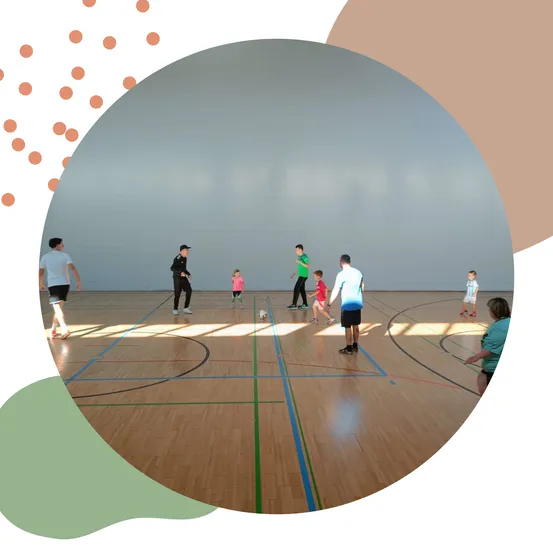 Eine Gruppe von Kindern und Erwachsenen spielt Fußball auf einem Hallenplatz. Einige gehen, während andere auf den Ball zulaufen.