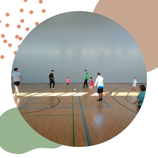 Eine Gruppe von Kindern und Erwachsenen spielt Fußball auf einem Hallenplatz. Einige gehen, während andere auf den Ball zulaufen.