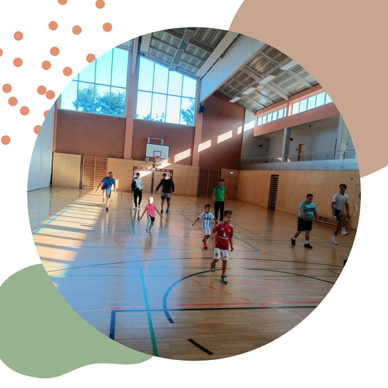 Kinder spielen eine Partie Fußball in einer Turnhalle mit Holzböden und Glasfenstern.