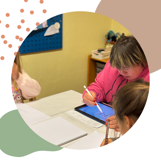 Zwei Kinder sitzen an einem Tisch, eines konzentriert sich auf ein Tablet, das andere hat einen Stift. Sie befinden sich in einem Klassenzimmer mit Papier und einem Bulletin Board im Blickfeld.