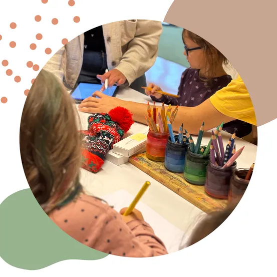 Kinder zeichnen und bunte Stifte sind auf dem Tisch. Ein Mädchen hält ein Tablet. Eine Lehrerin hilft ihr.