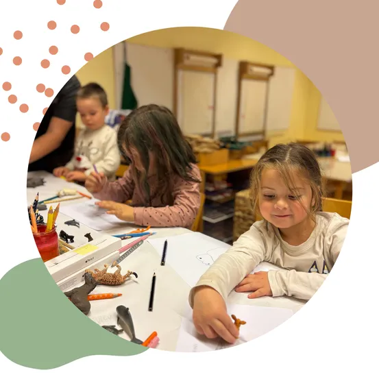 Drei Kinder sitzen an einem Tisch und sind in einer Aktivität vertieft. Ein Mädchen schaut sich einen Spielzeug-Dinosaurier an, während ein anderes Mädchen mit Buntstiften zeichnet. Ein Junge sitzt neben ihnen.