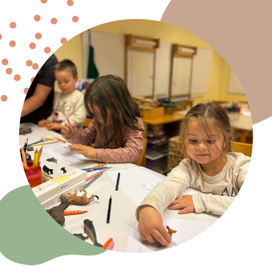 Drei Kinder sitzen an einem Tisch und sind in einer Aktivität vertieft. Ein Mädchen schaut sich einen Spielzeug-Dinosaurier an, während ein anderes Mädchen mit Buntstiften zeichnet. Ein Junge sitzt neben ihnen.
