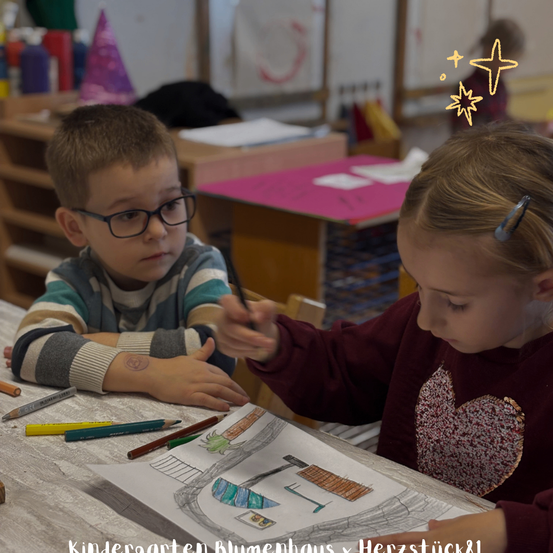 Zwei Kinder in einem Klassenzimmer zeichnen auf einem Papier, eines mit Brille, das andere mit einem Herz auf dem Shirt. Dahinter sind Tische mit Papieren und ein Regal mit Flaschen.