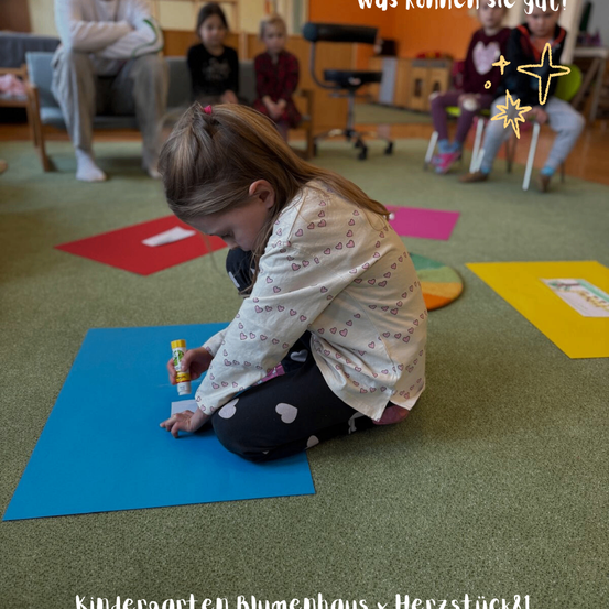Ein Mädchen sitzt auf dem Boden in einem Kindergartenraum und trägt Kleber auf ein blaues Papier auf. Andere Kinder sitzen auf Stühlen im Hintergrund. Der Boden hat bunte Matten. Ein Stern ist über ihr gezeichnet.