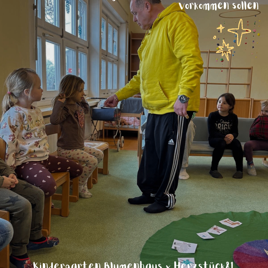 Ein Mann in einer gelben Jacke führt eine Gruppe von Kindern in einem Klassenzimmer an. Die Kinder sind sitzend und beobachten ihn. Das Klassenzimmer hat Teppich und Spielzeug auf dem Boden.