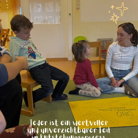 Eine Gruppe von Kindern und eine erwachsene Frau sitzen auf dem Boden, möglicherweise in einem Klassenzimmer. Die Frau lächelt und schaut ein Mädchen an. Das Mädchen schaut einen Jungen an. Sie sind alle in eine Diskussion verwickelt. Der Junge trägt ein grünes Shirt mit einem Cartoon-Charakter. Das Mädchen trägt ein rotes Langarmshirt.