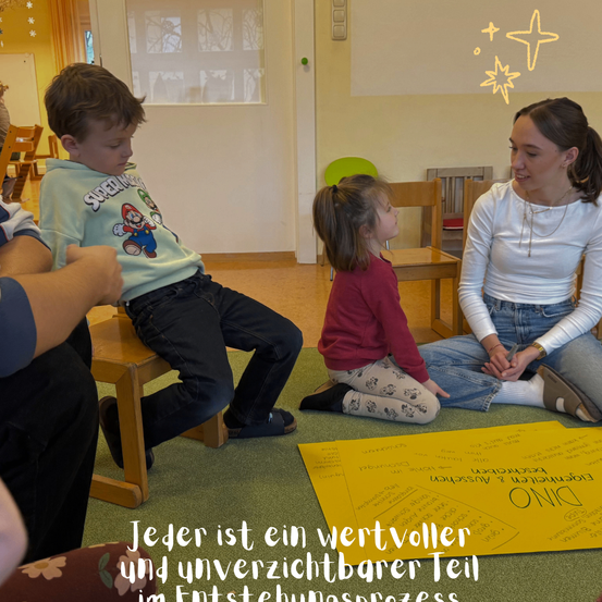 Eine Gruppe von Kindern und eine erwachsene Frau sitzen auf dem Boden, möglicherweise in einem Klassenzimmer. Die Frau lächelt und schaut ein Mädchen an. Das Mädchen schaut einen Jungen an. Sie sind alle in eine Diskussion verwickelt. Der Junge trägt ein grünes Shirt mit einem Cartoon-Charakter. Das Mädchen trägt ein rotes Langarmshirt.