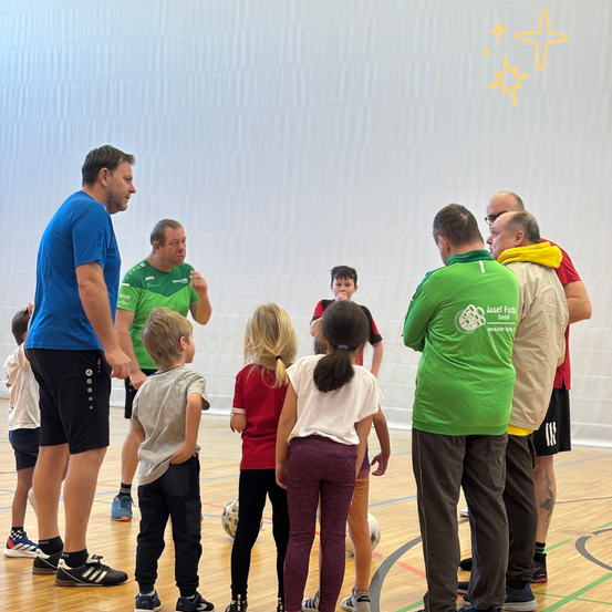 Eine Gruppe von Kindern und Erwachsenen in einer Turnhalle, einige in Sporttrikots. Ein Mann in einem blauen T-Shirt spricht mit einem anderen Mann, während Kinder um einen Fußball herumstehen.