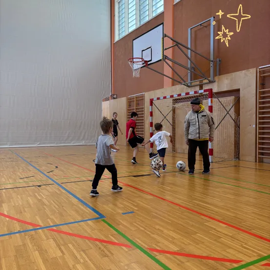 In einer Turnhalle spielen Kinder Fußball, während ein Mann zuschaut. Der Platz hat Basketballkörbe und Holzböden mit Markierungen.