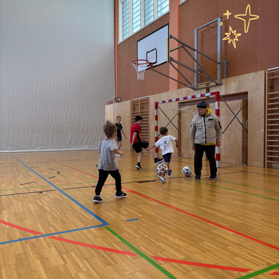 In einer Turnhalle spielen Kinder Fußball, während ein Mann zuschaut. Der Platz hat Basketballkörbe und Holzböden mit Markierungen.