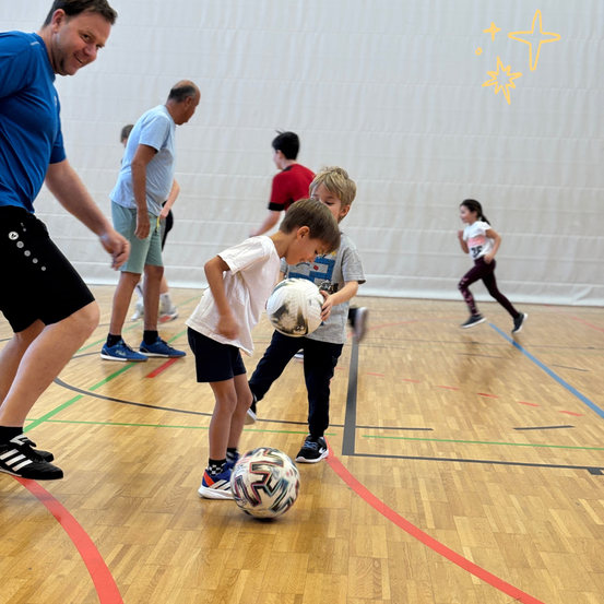 Eine Gruppe von Kindern spielt drinnen Fußball. Ein Junge hält den Ball und sieht nach unten.