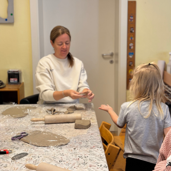 Eine Frau steht mit einem Kind an einem Tisch und beschäftigt sich mit Tonarbeiten. Die Frau hält ein Stück Ton, während das Kind zuschaut. Verschiedene Tonwerkzeuge und Gegenstände sind auf dem Tisch.