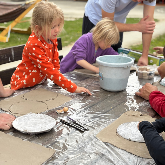 Mehrere Kinder stellen auf einem Tisch Tonkreise her, angeleitet von Erwachsenen. Sie benutzen Werkzeuge und haben Eimer in der Nähe. Der Ort scheint im Freien zu sein, mit einem Rasenbereich.