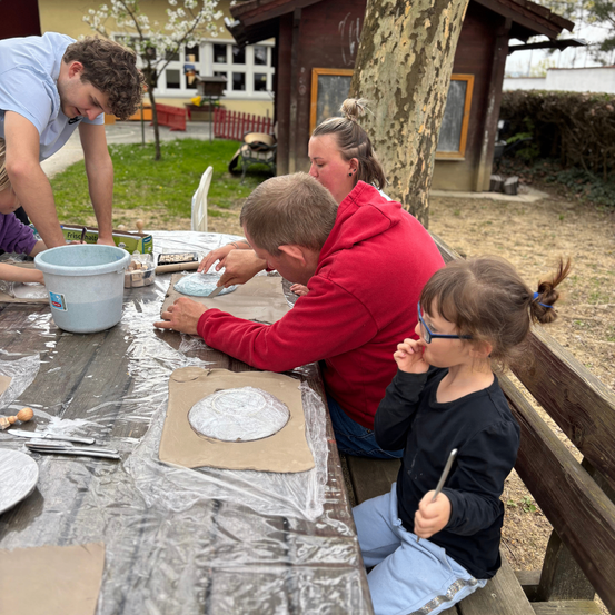 Eine Familie arbeitet draußen an einem Tisch an Töpferwaren in der Nähe eines Hauses. Ein Erwachsener führt ein Kind an. Ein anderes Kind beobachtet und ein Mann hält einen Eimer.