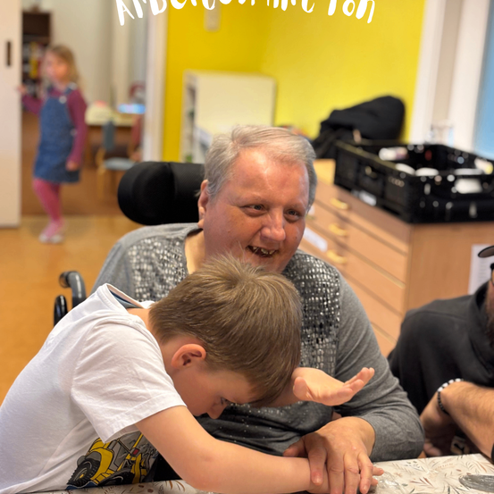Ein älterer Mann im Rollstuhl lächelt, während er mit einem jungen Jungen interagiert. Ein Mädchen im Hintergrund schaut zu ihnen.