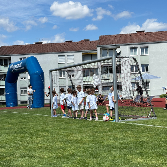 Eine Gruppe von Kindern in weißen Shirts steht in der Nähe eines Fußballtors auf einem Feld. Dahinter befindet sich ein blauer Bogen, ein Gebäude mit mehreren Fenstern und eine Person, die ein Kind hält.