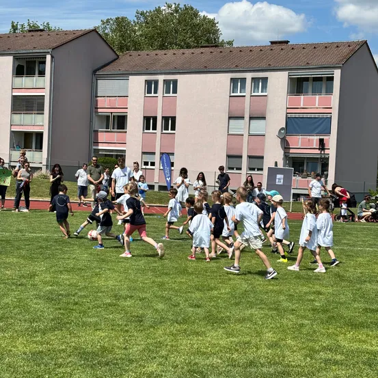Mehrere Kinder rennen im Kreis auf einem Rasenfeld, einige halten Fußballbälle. Im Hintergrund stehen oder gehen mehrere Erwachsene in der Nähe von zwei Wohngebäuden.