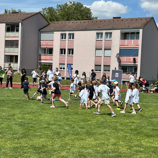 Mehrere Kinder rennen im Kreis auf einem Rasenfeld, einige halten Fußballbälle. Im Hintergrund stehen oder gehen mehrere Erwachsene in der Nähe von zwei Wohngebäuden.