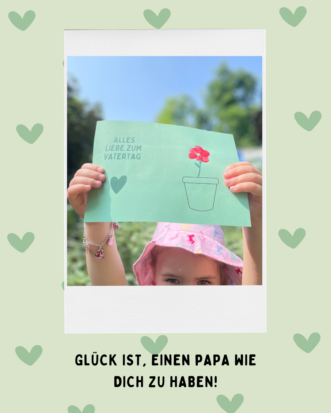 Ein Kind hält eine Karte mit einer Blume und dem Text 'Alles Liebe zum Vatertag' im Garten. Die Karte ist grün mit einer roten Blume und den Worten 'Alles Liebe zum Vatertag'. Das Kind trägt einen rosa Hut und eine Armband.