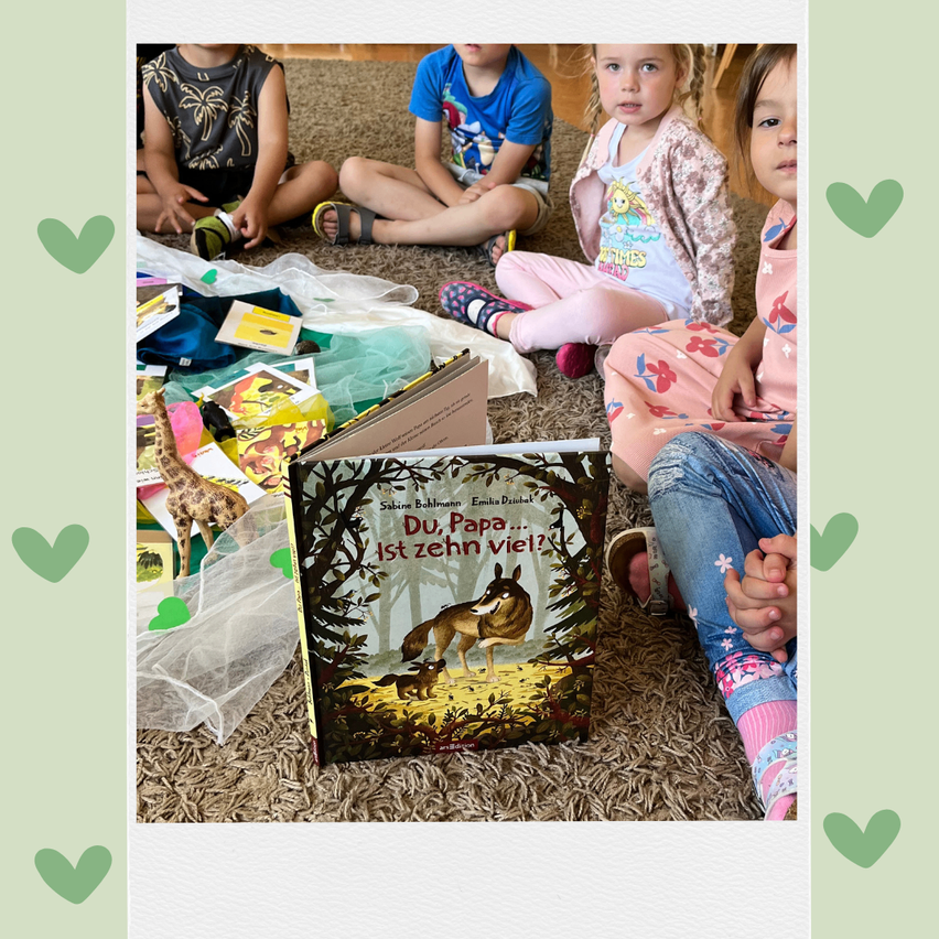Vier Kinder sitzen mit Spielzeug und einem Buch mit dem Titel Du, Papa ist zehn viel? auf dem Boden. Sie scheinen gemeinsam zu lesen oder zu spielen.