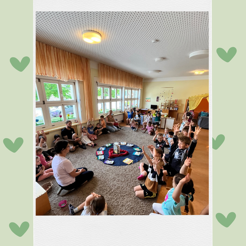 Eine Gruppe von Kindern und Erwachsenen sitzt im Kreis auf dem Boden. Eine Frau in einem weißen T-Shirt leitet die Gruppe. Der Raum hat eine weiße Decke mit Lampen und eine gelbe Wand.