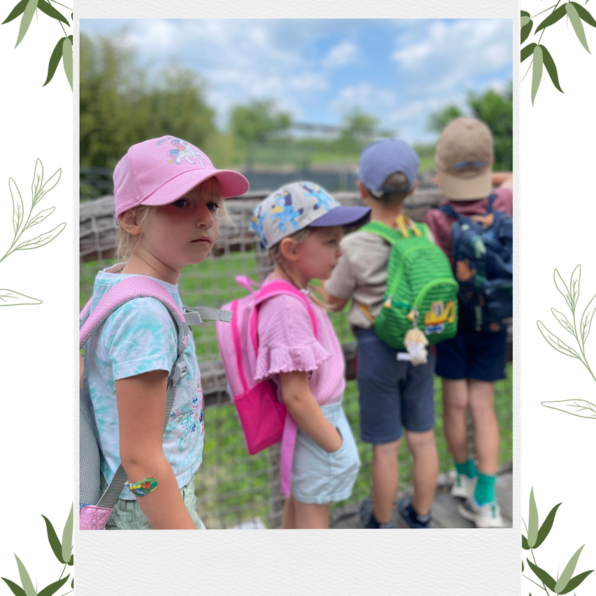 Eine Gruppe von Kindern, alle mit Mützen und Rucksäcken, stehen draußen in der Nähe eines Zauns. Die Kinder schauen alle in die gleiche Richtung.