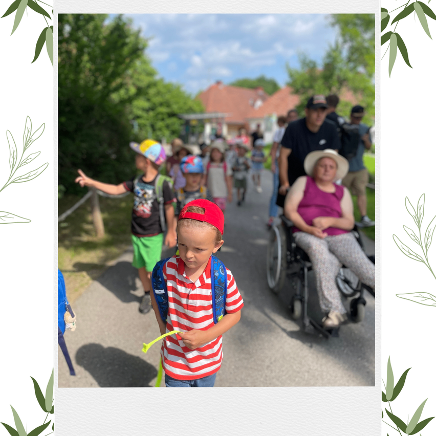 Eine Gruppe von Kindern in Hüten, die auf einem Pfad gehen, mit einem Mann in einem Rollstuhl in der Nähe. Im Hintergrund befindet sich ein Gebäude und Bäume.