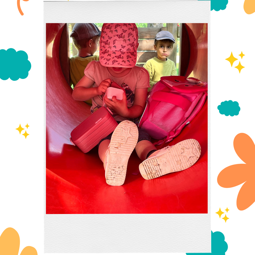 Ein kleines Mädchen sitzt in einem roten Tunnel und hält ein Handy, während zwei Jungen hinter ihr stehen, einer mit Hut und der andere mit Rucksack.