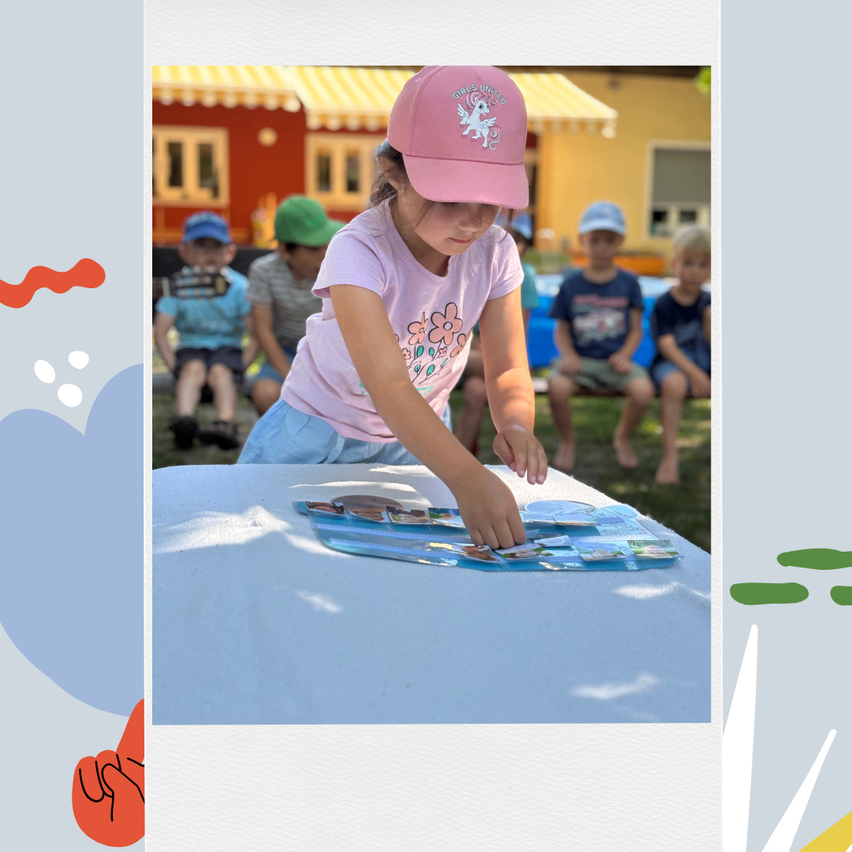 Ein junges Mädchen in einer rosa Mütze arbeitet draußen am Tisch an einem Puzzle. Hinter ihr sitzen Kinder auf einer Bank. Gebäude sind im Hintergrund.