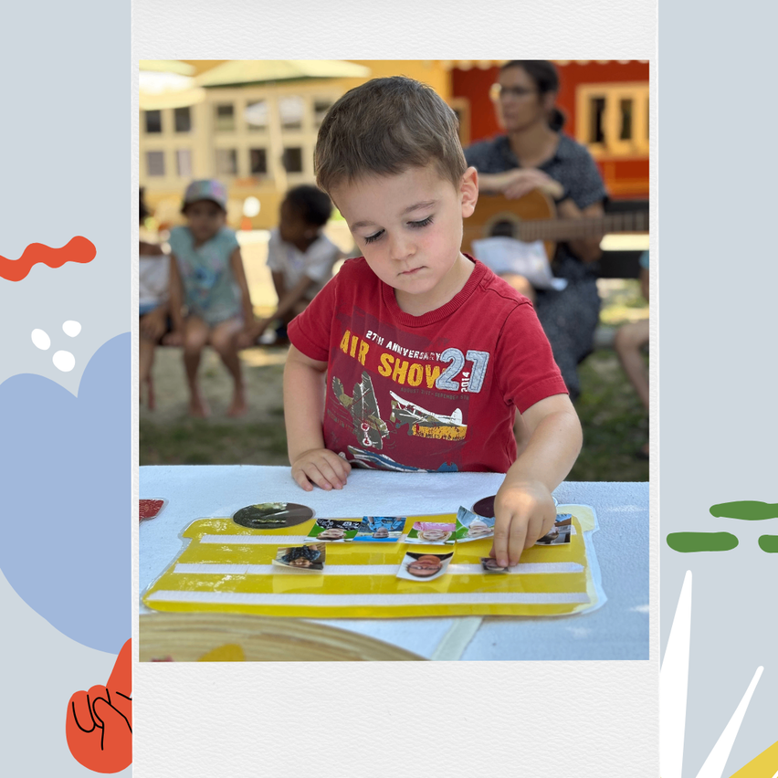 Ein kleiner Junge spielt ein Spiel mit Bildern auf einem gelben Tablett, mit anderen Kindern und einem Erwachsenen in der Nähe.