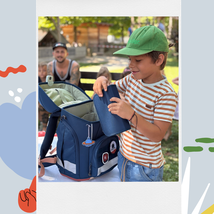 Ein Junge mit grüner Mütze und gestreiftem Shirt öffnet einen blauen Rucksack mit UFO-Aufkleber in einem Park.