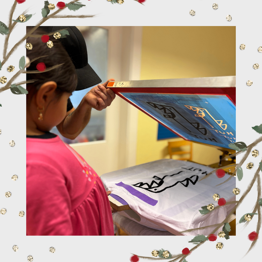 Ein Kind in einem rosa T-Shirt hält eine Siebdruckplatte, während eine Person in einem schwarzen Hut hilft. Ein weißes T-Shirt liegt auf einem Tisch, der mit einem weißen Tuch bedeckt ist und von dekorativen Elementen umgeben ist.