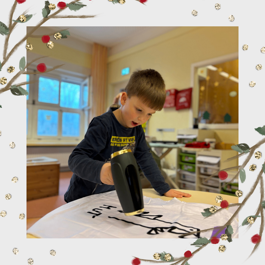 Ein junger Junge arbeitet an einem weißen Stoff mit einer schwarzen Zeichnung und hält eine Heißklebepistole. Er befindet sich in einem Raum mit Fenster und Schränken.