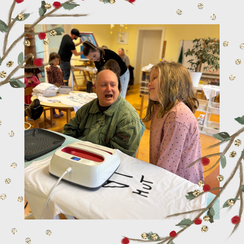 Ein Mann lacht, während ein Mädchen zusieht, beide in einem Raum mit einem Tisch und einem Heißpressgerät. Im Hintergrund sind Menschen.