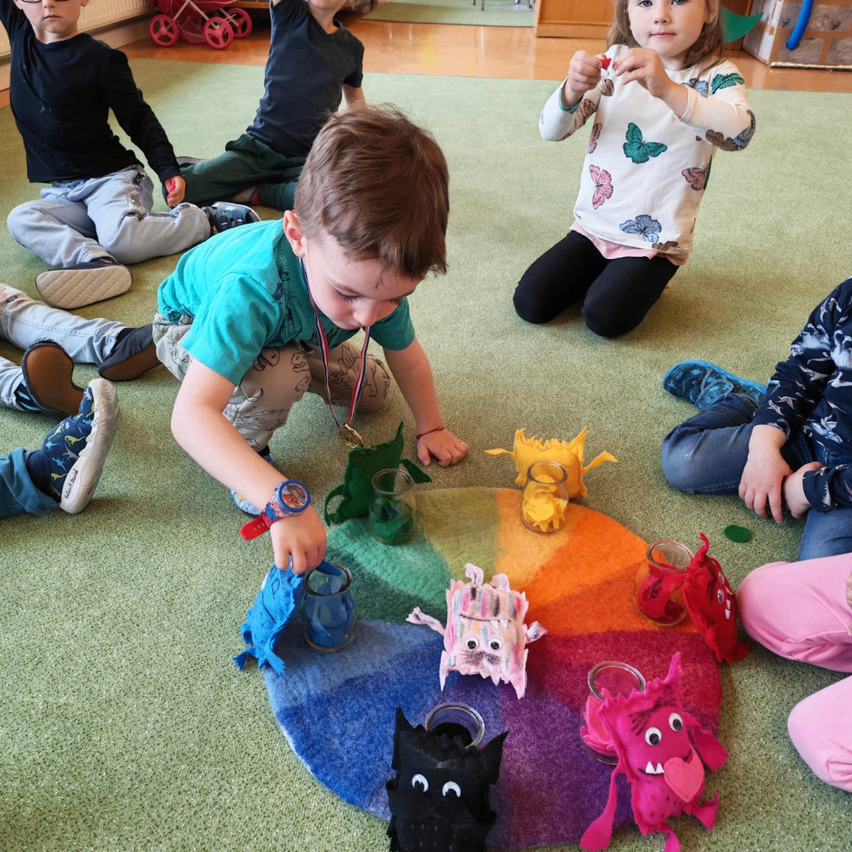 Eine Gruppe von Kindern sitzt auf dem Boden und spielt mit einem kreisförmigen Teppich, auf dem verschiedene bunte Spielzeuge liegen. Jedes Kind ist mit einem anderen Spielzeug beschäftigt.