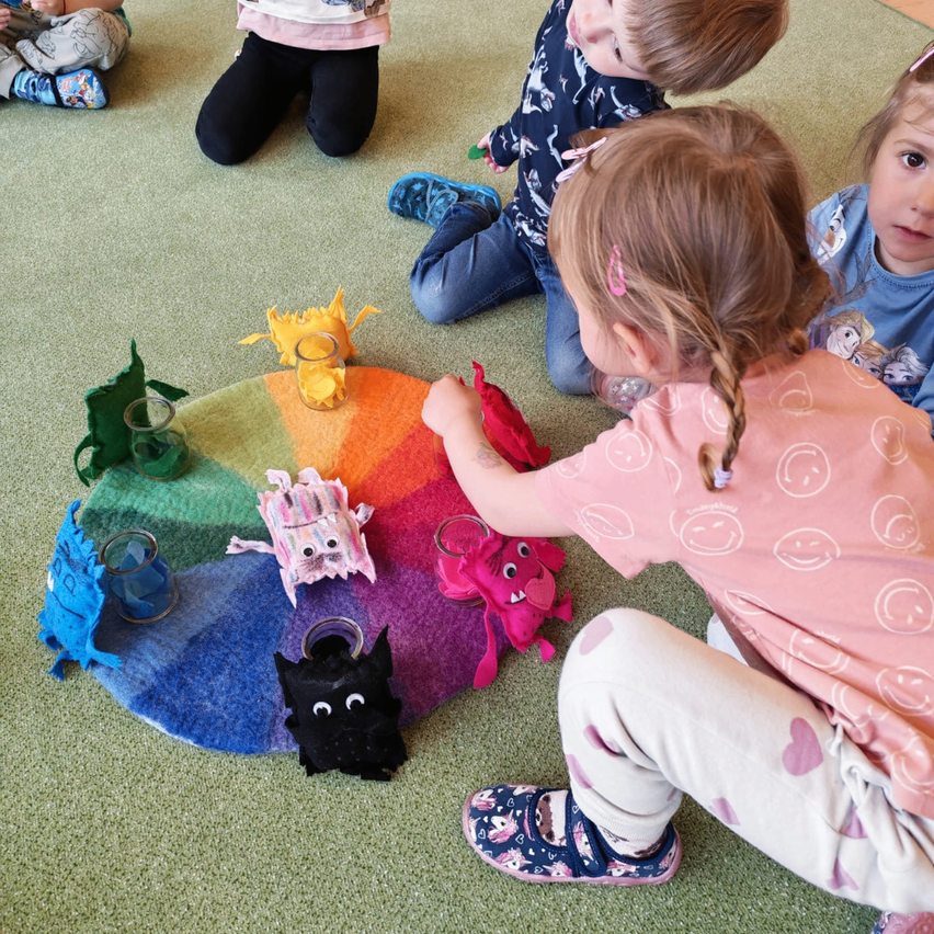Vier Kinder sitzen um eine bunte Matte mit verschiedenen Plüschtieren herum. Ein Mädchen in einem rosa Shirt greift nach einem schwarzen Plüschhund.