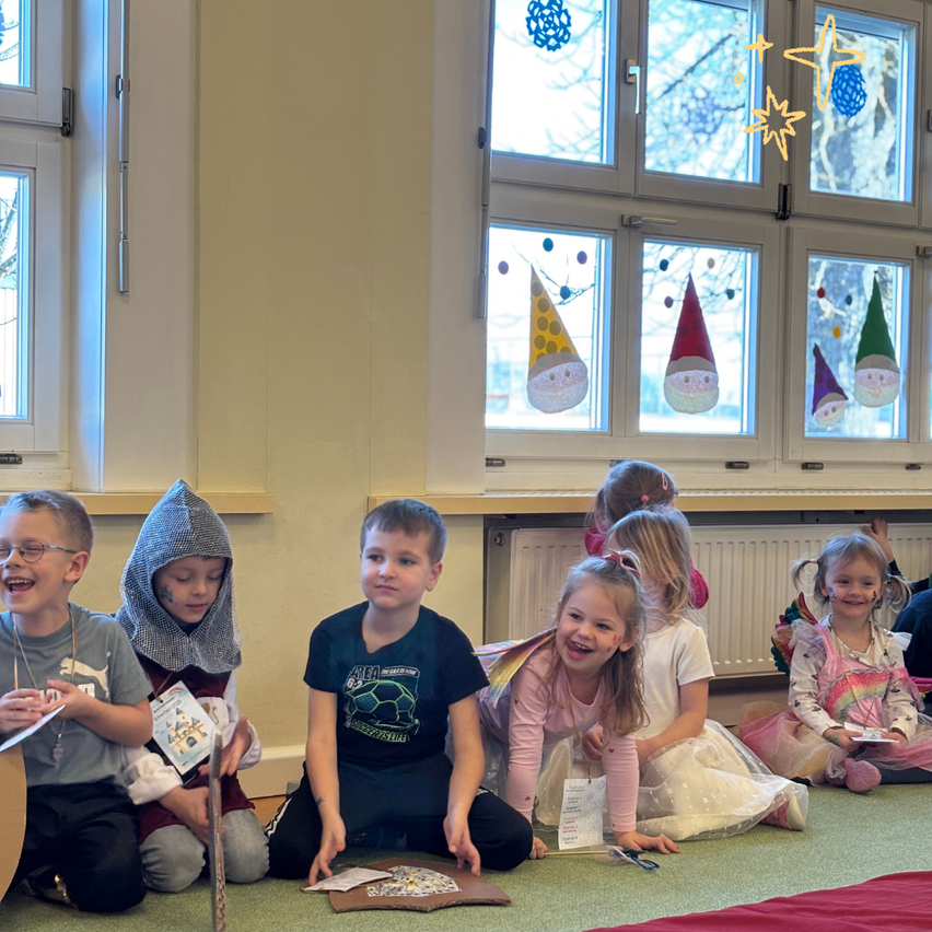 Mehrere Kinder sitzen auf dem Boden, einige in Kostümen, lächeln und sind in einer Aktivität verwickelt. Ein Heizkörper und dekorierte Fenster sind im Hintergrund zu sehen.