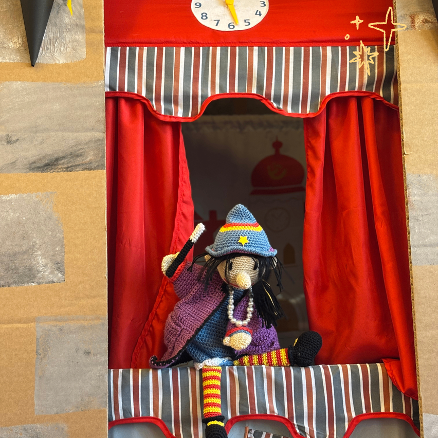 Eine Puppe in einer Hexen-Kostüm sitzt in einem kleinen Papp-Marionettentheater. Das Theater hat rote Vorhänge, eine Uhr darüber und ein Sterndesign.