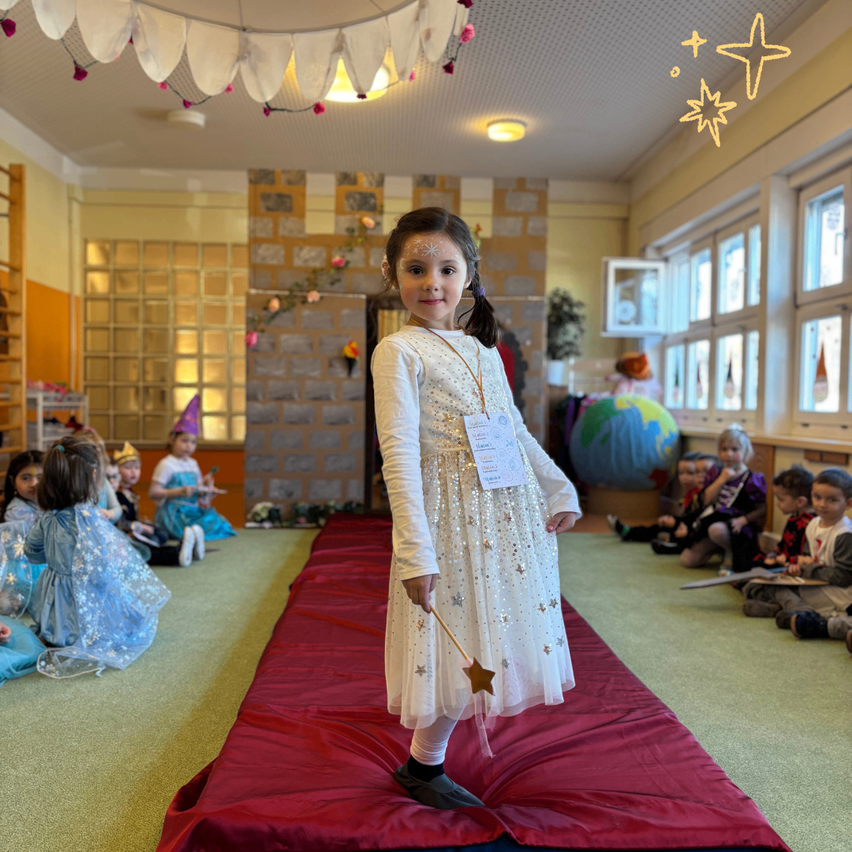 Ein kleines Mädchen steht auf einem roten Teppich und hält einen Sternstab, während andere Kinder um sie herum auf dem Boden sitzen. Der Raum ist mit Sternen und Lichterketten dekoriert.