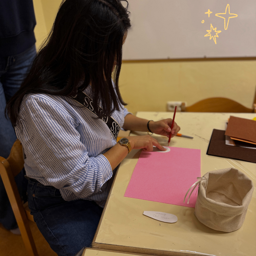 Eine Frau sitzt an einem Schreibtisch und arbeitet mit einem roten Stift an einem rosa Papier. Sie trägt ein gestreiftes Hemd und Jeans. Ein Beutel steht auf dem Tisch. Ein Whiteboard ist im Hintergrund.