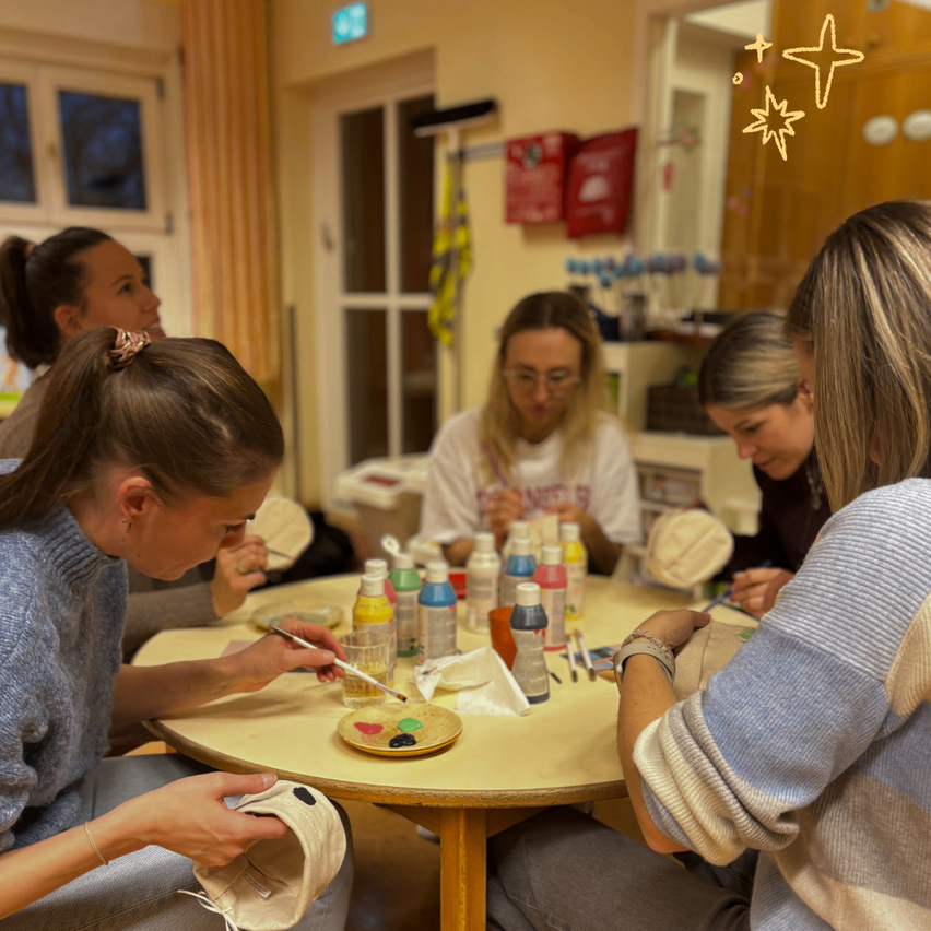 Eine Gruppe von Frauen sitzt um einen Tisch herum und malt auf Hüte. Es gibt verschiedene Flaschen mit Farbe und einen Malkasten auf dem Tisch. Der Raum hat ein Fenster mit Vorhängen und einen Feuerlöscher an der Wand.