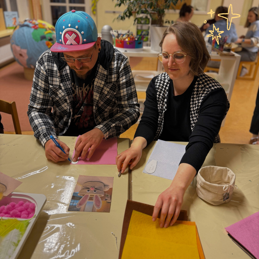 Ein Mann und eine Frau sitzen an einem Tisch und basteln. Sie sind von verschiedenen Bastelmaterialien umgeben.