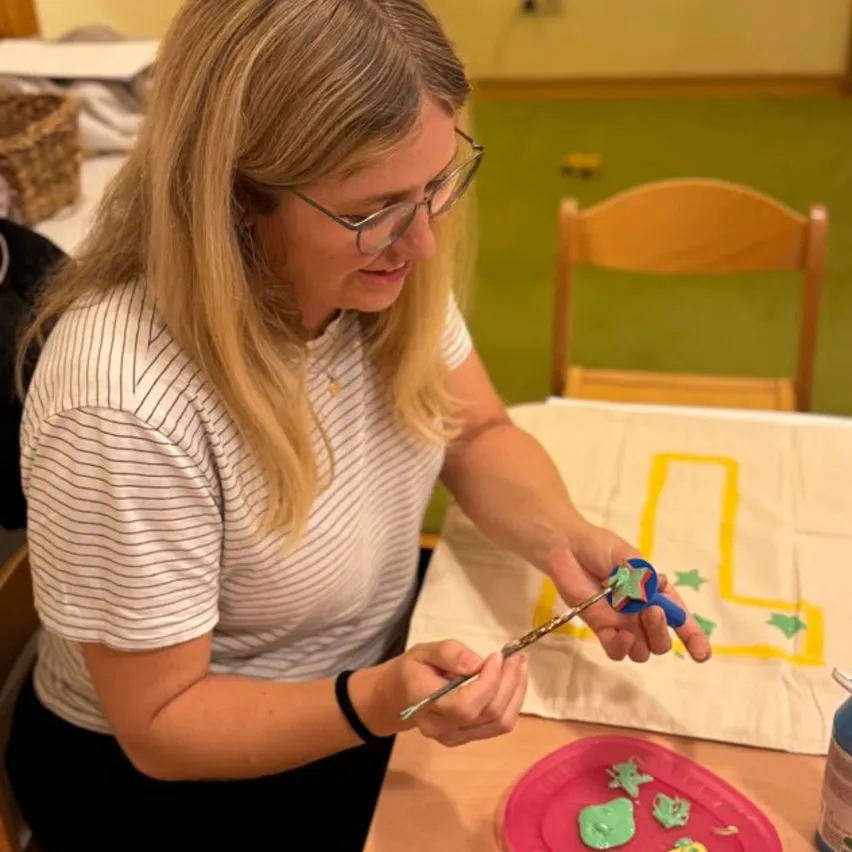 Eine Frau mit Brille und gestreiftem Shirt malt ein Bastelprojekt mit einem Pinsel auf einem Tisch.