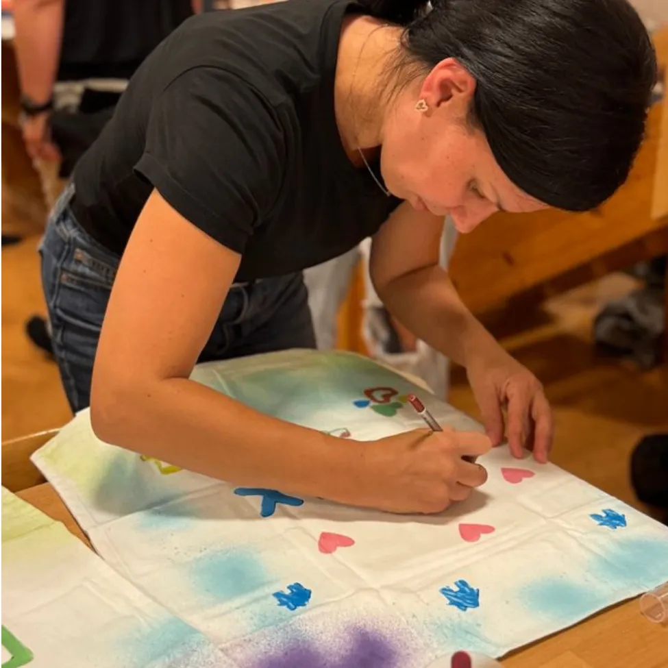 Eine Frau in einem schwarzen T-Shirt und Jeans malt ein weißes Tuch mit bunten Designs. Sie konzentriert sich auf ihre Arbeit, mit einigen anderen Personen im Hintergrund.