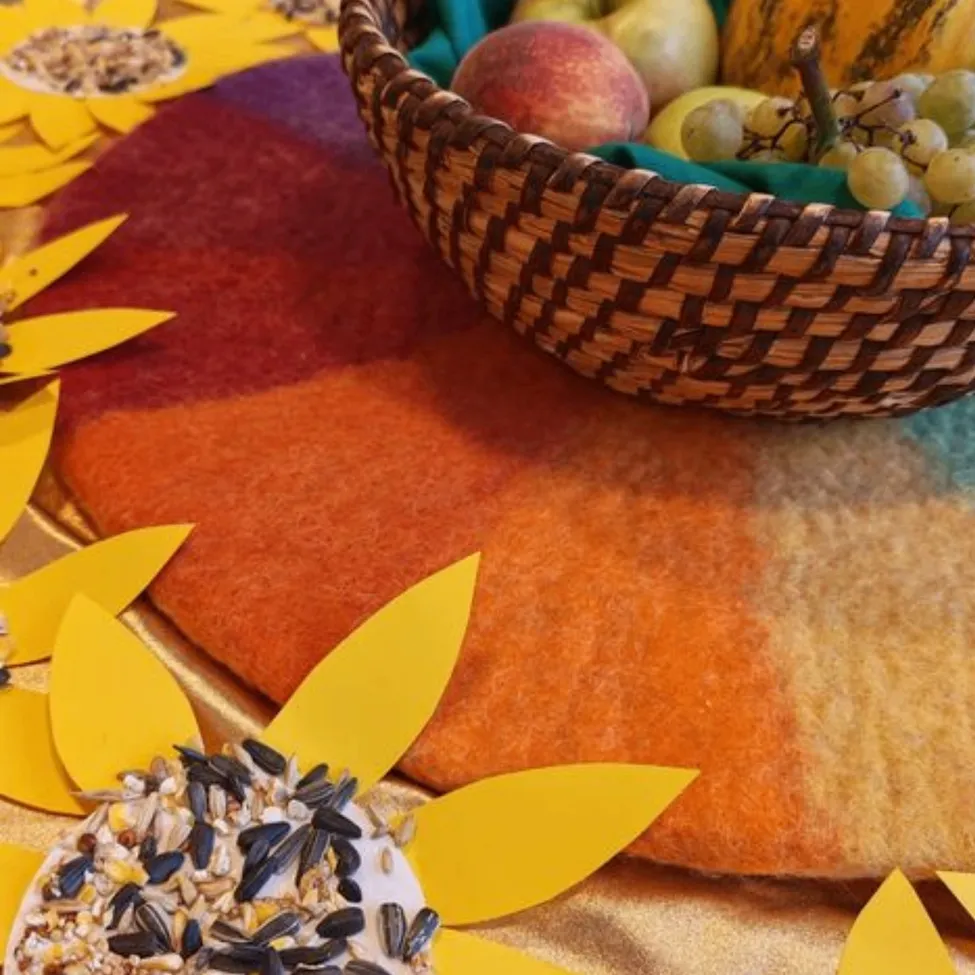 Ein Korb mit Früchten wie Äpfeln, Trauben und einer Wassermelone steht auf einem farbenfrohen Filzuntersetzer mit Sonnenblumendesign.