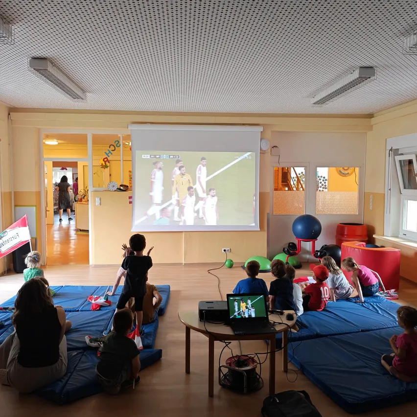 Eine Gruppe von Kindern und Erwachsenen schaut sich ein Fußballspiel auf einer Projektionsleinwand in einem Raum an. Blaue Matten sind auf dem Boden verteilt.