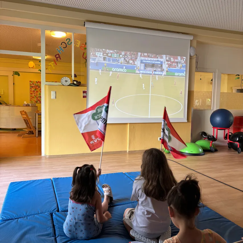 Drei Mädchen sitzen auf blauen Kissen und sehen sich ein Fußballspiel auf einer Projektionsleinwand in einem gelben Raum an. Zwei halten Flaggen, eine mit einem rot-weißen Design.